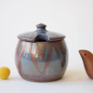 May include: A small, round, ceramic sugar bowl with a lid. The bowl is decorated with a blue, brown, and copper glaze. The lid has a small spout for pouring.