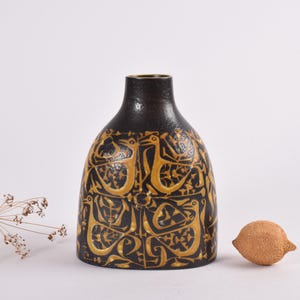 Puede incluir: Un jarrón de cerámica negro con un patrón geométrico dorado. El jarrón tiene una base ancha y un cuello estrecho.