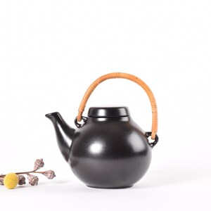 Op de afbeelding: Een zwarte keramische theepot met een afgeronde body en een gebogen tuit. Hij heeft een geweven rotan handvat en een bijpassende zwarte deksel. Een takje met gele bloemen en gedroogde zaaddozen staat op de voorgrond.