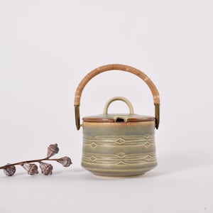 Può includere: Un vaso di ceramica con coperchio e manico intrecciato. Il vaso è verde chiaro con un motivo a rombi al centro. Il coperchio è marrone e il manico è marrone chiaro. Un rametto di fiori secchi è a sinistra.