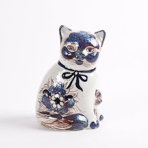 May include: A white ceramic cat figurine with blue and brown painted details. The cat is sitting with its head tilted slightly to the side. The cat has a blue and white ribbon around its neck and a floral design on its side.