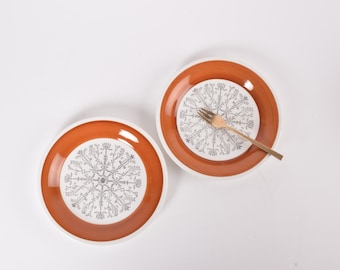 May include: Two ceramic plates with a white interior and a rust-colored rim. Each plate features a detailed, symmetrical black and white floral design. One plate has a gold fork resting on it. The plates are on a white surface.