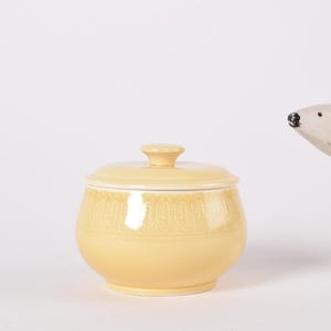 May include: A yellow ceramic sugar bowl with a lid and a decorative pattern. The bowl is round and has a smooth, glossy finish.