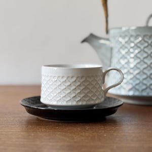 以下が含まれることがあります： ハート柄の白いセラミック製のティーカップとソーサー。ティーカップにはハンドルが付いており、ソーサーは黒色です。背景にはハート柄のグレーのセラミック製のティーポットがあります。