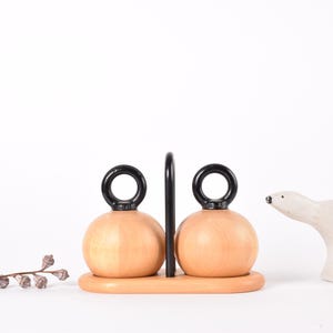 Może przedstawiać: Drewniany zestaw solniczki i pieprzniczki z czarnym uchwytem i czarnymi uchwytami w kształcie pierścieni. Zestaw stoi na drewnianej podstawie. Po prawej stronie zestawu znajduje się biała figurka niedźwiedzia polarnego. Po lewej stronie znajduje się mała gałązka.