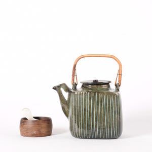 Peut inclure: Une théière en céramique rayée verte et brune avec une poignée en bambou. La théière a un couvercle et un bec verseur. Il y a un petit bol en bois avec une cuillère blanche à côté de la théière.