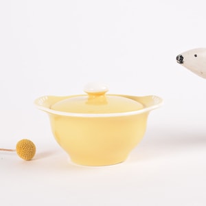 以下が含まれることがあります： 白い蓋と白い縁のある黄色のセラミック製の砂糖入れ。ボウルは白い表面の上に置かれています