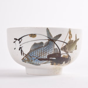 Puede incluir: Cuenco de cerámica blanca con base, decorado con un diseño de peces azules y marrones. El cuenco presenta una ilustración detallada de peces y plantas acuáticas, con detalles en negro y marrón. El cuenco es adecuado para servir alimentos.