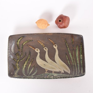 May include: A rectangular ceramic serving dish with a brown glaze. The dish features a painted design of three white birds with long necks and beaks, standing in a field of green reeds.