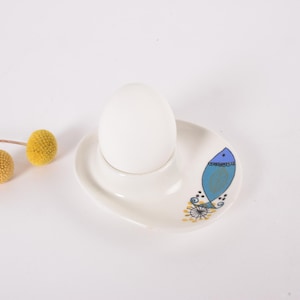 Può includere: Un portauovo in ceramica bianca con un disegno di pesce blu e un uovo bianco. Due boccioli di fiori gialli sono a sinistra del portauovo. Il portauovo è su una superficie bianca.