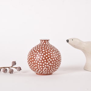 May include: A brown ceramic vase with white polka dots, a white rim, and a small wooden polar bear figurine. A sprig of dried seed pods is on the left. The objects are on a white background.