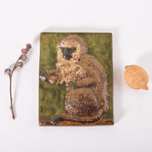 Puede incluir: Una baldosa de cerámica rectangular con un relieve de un mono en tonos marrones y beige sobre un fondo verde. También hay un pequeño objeto texturizado de color beige y un tallo de planta seca.