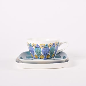 Può includere: Un set di tazza e piattino bianchi con un motivo a pesci blu, verde acqua e gialli. La tazza ha un manico e poggia su un piattino abbinato e un piatto rettangolare. Il set è su sfondo bianco.