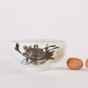 Puede incluir: Un pequeño cuenco de cerámica blanca con un diseño de pez en marrón y verde. El cuenco tiene forma redondeada y un pie pequeño. Dos nueces marrones están junto al cuenco.