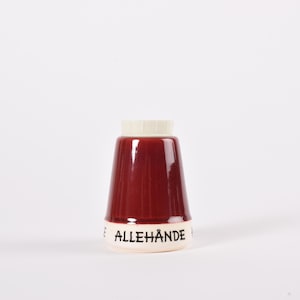Puede incluir: Un salero de cerámica granate con base y tapa blancas. La palabra "ALLEHÄNDE" está impresa en negro en la base. El salero tiene forma cónica y un acabado brillante, adecuado para la cocina.