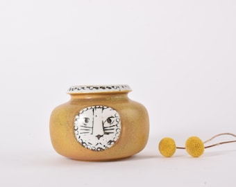 May include: A small, round, ceramic vase with a lion face design. The vase is a warm, mustard yellow color with a white and black rim. The lion's face is white with black details. Two yellow flower heads are to the right.
