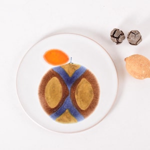 May include: A white ceramic plate with a stylized orange design. The orange is painted in shades of brown, blue, and yellow. The plate is on a white background.