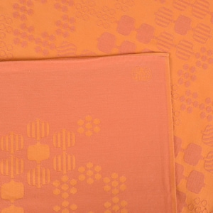 May include: A peach-coloured textile featuring a repeating pattern of geometric shapes in a lighter peach hue. The fabric is folded over, showing a solid peach colour on the underside. The design is abstract and decorative, suitable for home decor.