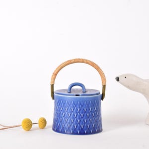 Puede incluir: Un azucarero de cerámica azul con un asa trenzada y un patrón geométrico. El tazón está sobre una superficie blanca.