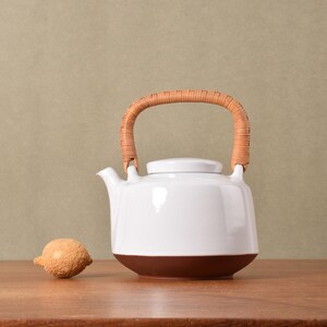 May include: A white ceramic teapot with a brown base and a curved, light brown woven handle. Beside the teapot sits a small, textured, light brown spherical object. The teapot features a short spout and a matching lid, creating a contrast.
