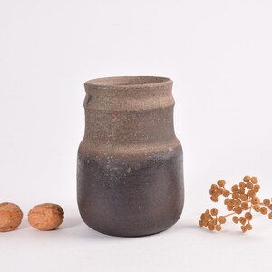 以下が含まれることがあります： 手作りの素朴なセラミック花瓶。濃い茶色のベースと、より明るく質感のある上部が特徴です。2つのクルミと乾燥した花の小枝が近くに配置されており、自然でアースカラーを基調としたテーマを連想させます。
