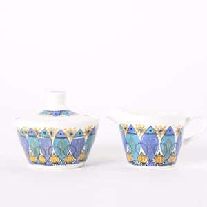 Può includere: Una zuccheriera in ceramica bianca con coperchio e una lattiera. Entrambi i pezzi presentano un motivo ripetuto di forme di foglie stilizzate blu, verde acqua e gialle. La zuccheriera ha un coperchio con una piccola maniglia.