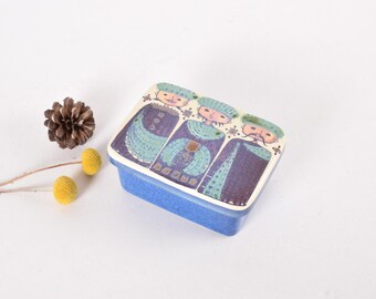 May include: A blue ceramic box with a white lid featuring a colorful illustration of three people wearing hats. The lid has a whimsical design with geometric patterns and stars.