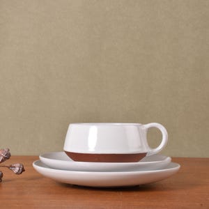 May include: A white ceramic cup with a brown base sits on a matching white saucer. The cup features a rounded body and a small handle. The saucer is circular with a smooth finish. The design is simple, featuring a two-tone color scheme.