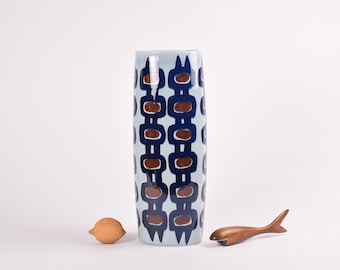 May include: A white ceramic vase with a blue and brown geometric pattern. The vase is tall and cylindrical.