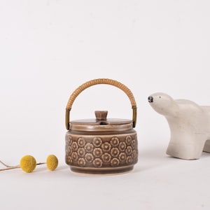 May include: A brown ceramic jar with a lid and a woven handle, decorated with a floral pattern. A small, off-white polar bear figurine is to the right. Two yellow, spherical flowers and a twig are in the foreground.