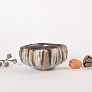 Puede incluir: Cuenco de cerámica hecho a mano con rayas verticales en tonos grises, marrones y crema. El cuenco tiene una forma irregular. Una nuez y vainas decorativas están junto al cuenco. El cuenco mide unos 15 cm de diámetro.