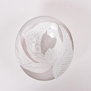 Puede incluir: Un cuenco de cerámica blanca con un diseño de una sirena con el pelo suelto y una cola que se asemeja a un ala de pájaro. El cuenco tiene un esmalte rosa claro.