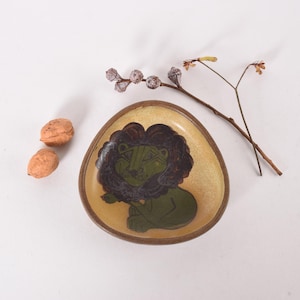 May include: A small, yellow ceramic dish with a brown rim. The dish features a green and brown illustration of a lion.
