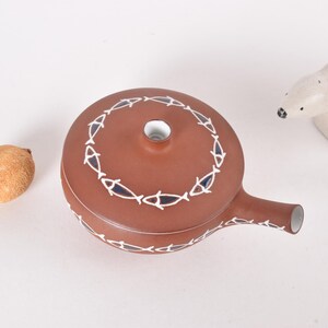 May include: A brown ceramic teapot with a white and blue fish design. The teapot has a spout and a lid.