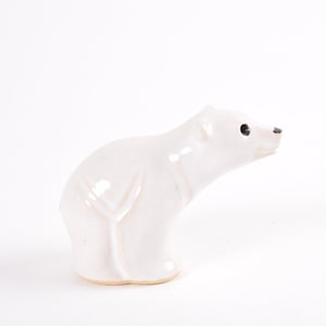 May include: White ceramic polar bear figurine. The bear is sitting on its haunches with its head turned to the side. The bear has black eyes and a small, pointed nose.
