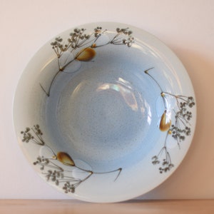 Può includere: Una ciotola in ceramica blu chiaro con un motivo floreale in marrone e giallo. La ciotola ha un bordo leggermente ondulato.