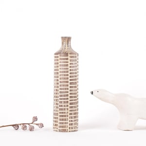 May include: A brown and white ceramic vase with a geometric pattern. The vase is tall and slender, and it is sitting on a white surface. A white ceramic polar bear figurine is also in the image.