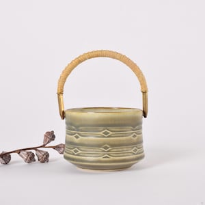 Op de afbeelding: Een kleine, olijfgroene keramische emmer met een geweven handvat en goudkleurige metalen accenten. De emmer heeft een herhalend ruitpatroon. Een decoratieve tak met kleine, gedroogde zaaddozen staat op de achtergrond.