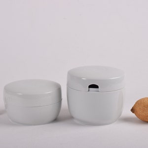 Puede incluir: Dos recipientes de plástico blanco con tapas. El recipiente más grande tiene una pequeña abertura en la tapa. El recipiente más pequeño tiene una tapa lisa. Ambos recipientes están sobre una superficie blanca.