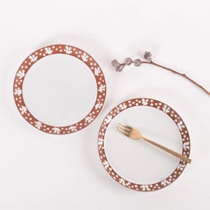 May include: Two white ceramic plates with a brown and white patterned border. The plates are on a white surface. A gold fork is on one of the plates.