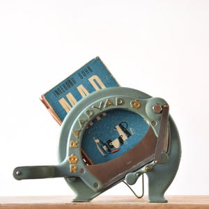 Puede incluir: Cortadora de pan vintage azul y dorada con una cuchilla curva. La cortadora tiene un mango y un libro con el texto "INGEBORG SUHR MAD" en su interior. Se encuentra sobre una superficie de madera.