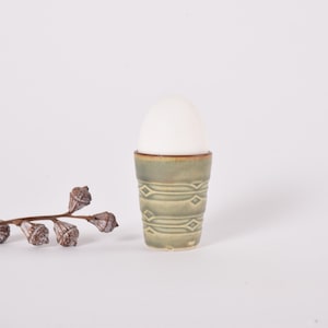 Puede incluir: Un huevero de cerámica verde claro con un diseño geométrico, que contiene un huevo blanco. Una ramita de eucalipto seco está a la izquierda del huevero, sobre un fondo blanco.