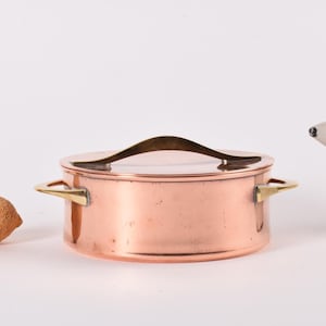 Può includere: Una casseruola di rame con coperchio e manici in ottone. Un limone di sughero e una statuina di orso polare bianco sono accanto alla pentola. La pentola è su sfondo bianco.