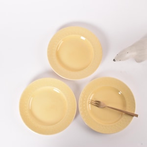May include: Three yellow ceramic plates with a decorative pattern around the edge. The plates are arranged in a triangle shape on a white surface.