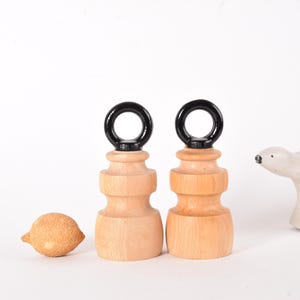 Może przedstawiać: Dwa drewniane uchwyty do ćwiczeń z czarnymi metalowymi pierścieniami. Uchwyty są wykonane z jasnego drewna i mają gładkie wykończenie.