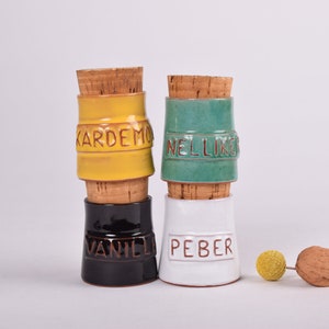 May include: Four ceramic spice jars with cork lids. The jars are stacked in pairs. The top jar is yellow with the word "KARDE MOM" written on it. The second jar is green with the word "NELLIK" written on it. The third jar is black with the word "VANILJ" written on it. The fourth jar is white with the word "PEBER" written on it.