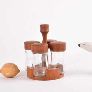Puede incluir: Un estante de especias de madera con cuatro tarros de vidrio y tapas de madera. El estante está hecho de madera oscura y tiene una base redonda. Los tarros son de vidrio transparente y tienen una forma ligeramente redondeada.