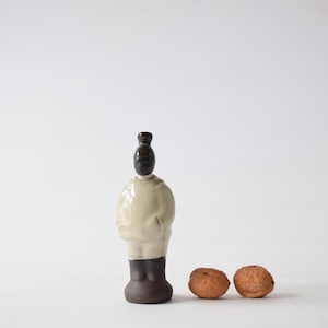 May include: A ceramic figurine of a person with a white body, brown head, and brown legs. The figurine is standing on a white surface with two brown walnuts in front of it.
