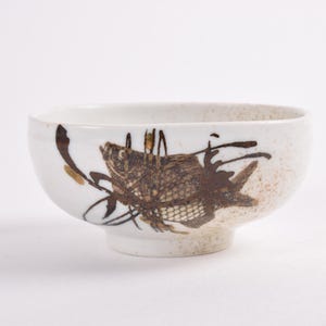 Puede incluir: Cuenco de cerámica blanca con una ilustración de pez marrón. El cuenco tiene forma redondeada y una pequeña base con pie. El diseño del pez es detallado con un aspecto texturizado y el cuenco tiene un acabado moteado.