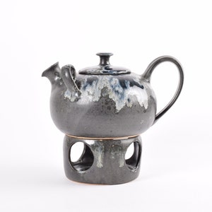 May include: A black ceramic teapot with a blue and white glaze sits on a matching stand with cutouts. The teapot has a round body and a small spout.